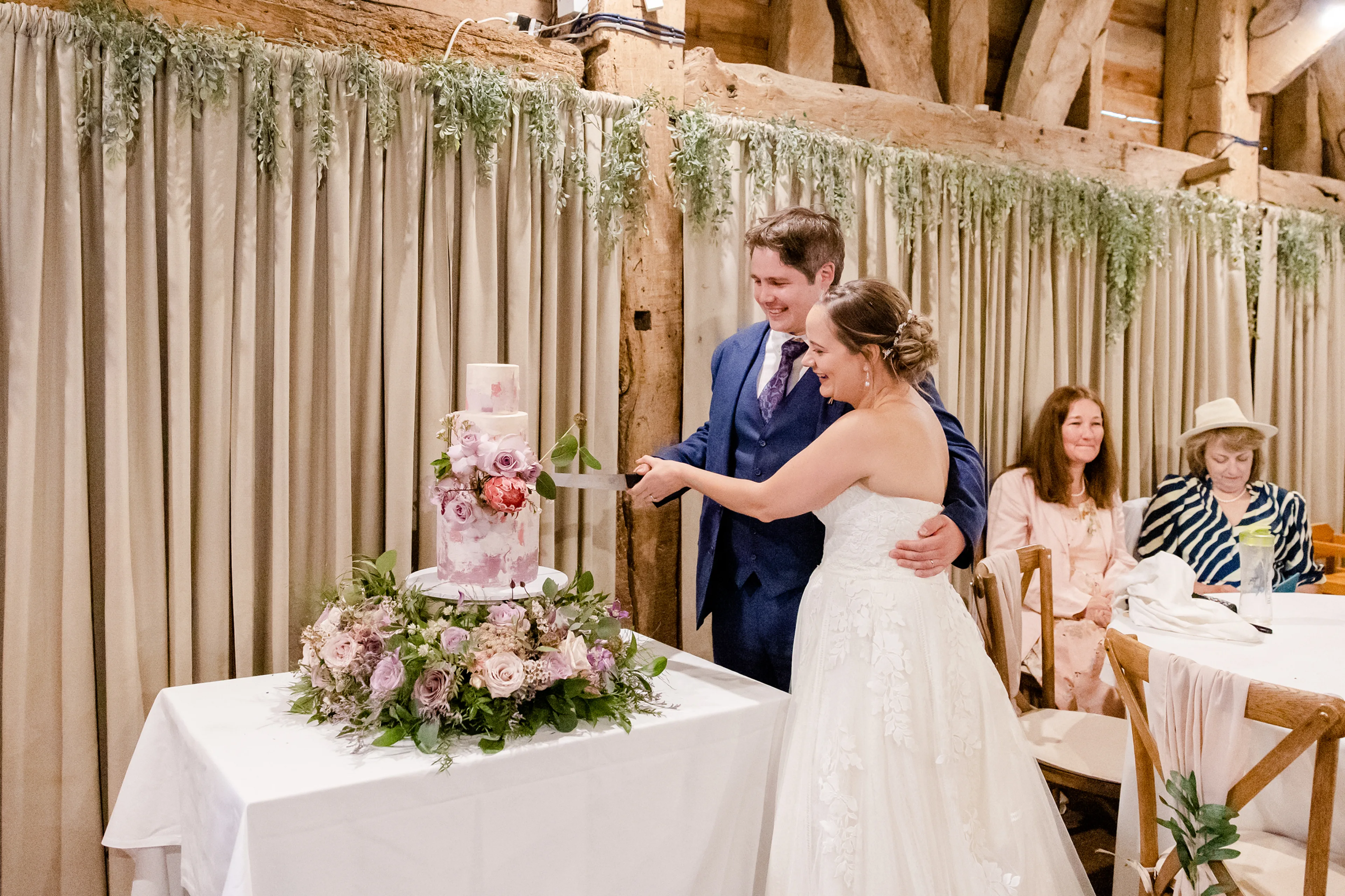 Couple Cutting Tree tier wedding cake in Mauve watercolour and floral arrangement with Protea flower and roses