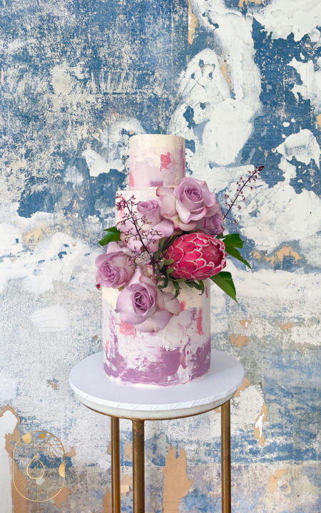 Tree tier wedding cake in Mauve watercolour and floral arrangement with Protea flower and roses. Colourful rustic background.